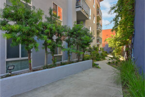 Outdoor walkway, above ground concrete planters with trees, balconies above walkway, surrounding landscaping